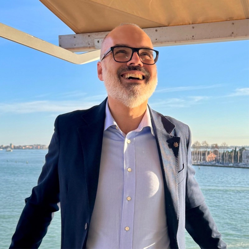 Portrait of Roberto Ongaro of Deserving Italy, laughing in a navy jacket on a boat, with a lagoon and a distant shoreline behind him.