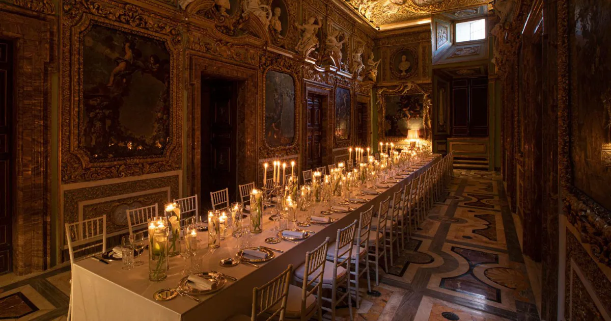 A long table set for dinner by candlelight in a frescoed Roman palace, with gilded stucco walls, painted panels and an inlaid marble floor.
