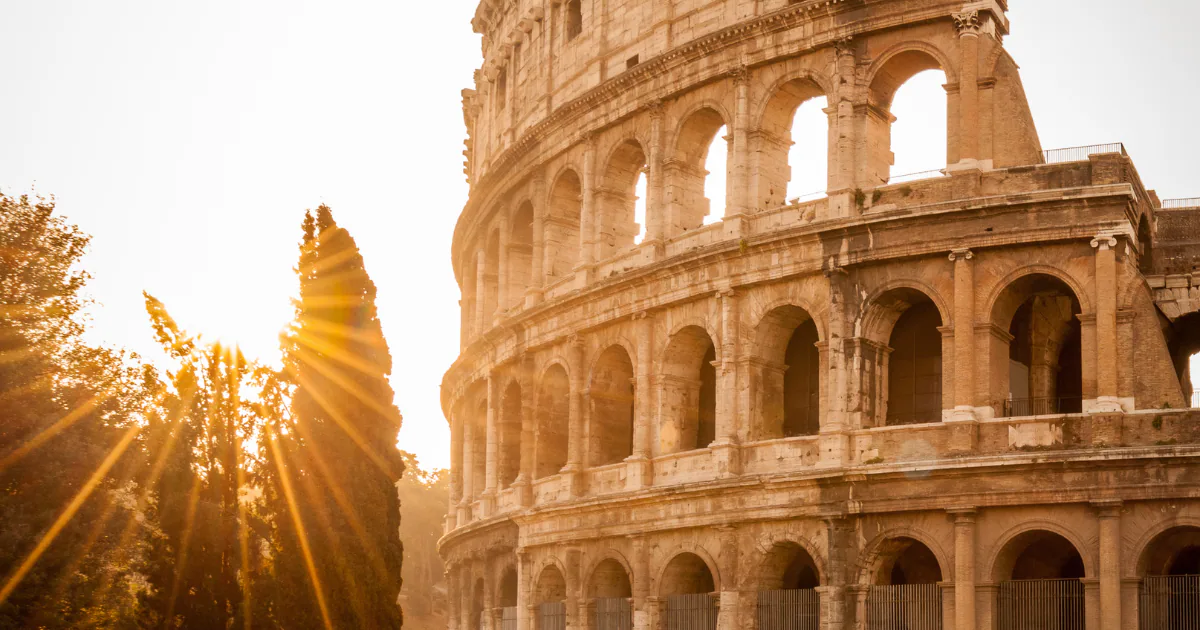 The tiered arches of the Colosseum in warm morning light, with sun rays breaking through the trees at its side.