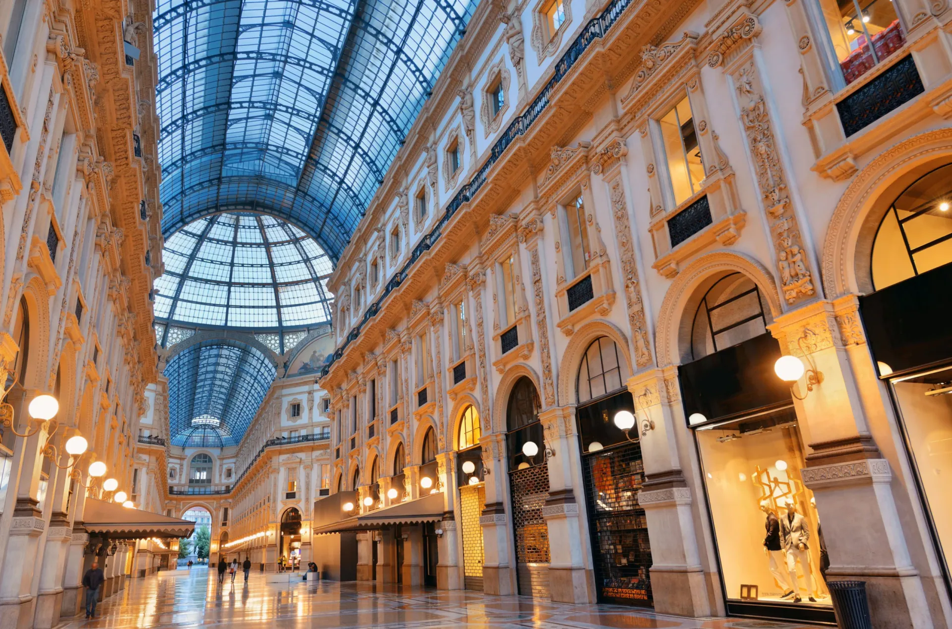 Low-angle perspective of the Galleria Vittorio Emanuele II in Milan, featuring its iconic glass and iron vaulted ceiling, ornate architectural details, and luxury shop windows glowing with warm light.