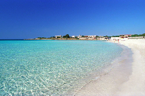 Shallow, transparent water over white sand on the Ionian coast of Salento, with low houses along the shoreline.
