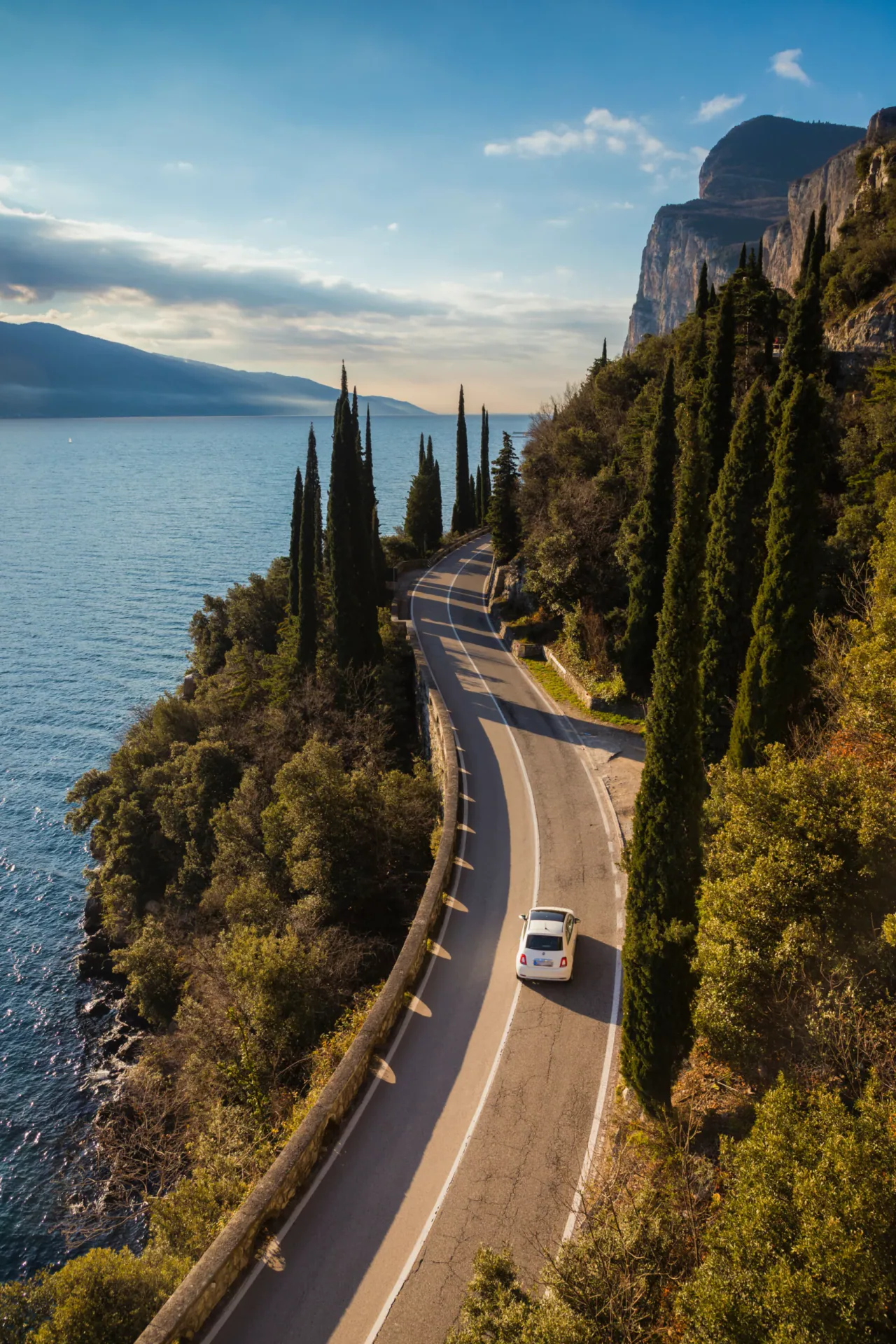 Aerial view of a white car driving on the winding Gardesana coastal road along Lake Garda, flanked by tall cypress trees and steep cliffs during sunset.