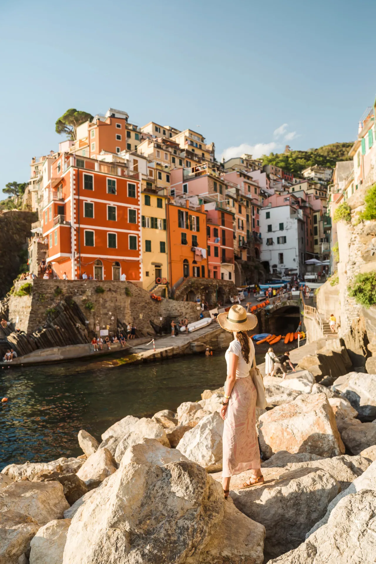 Woman wearing a straw hat and long skirt standing on the rocky breakwater, admiring the vibrant red and yellow houses of Riomaggiore village in the Cinque Terre, Italian Riviera, during sunset.
