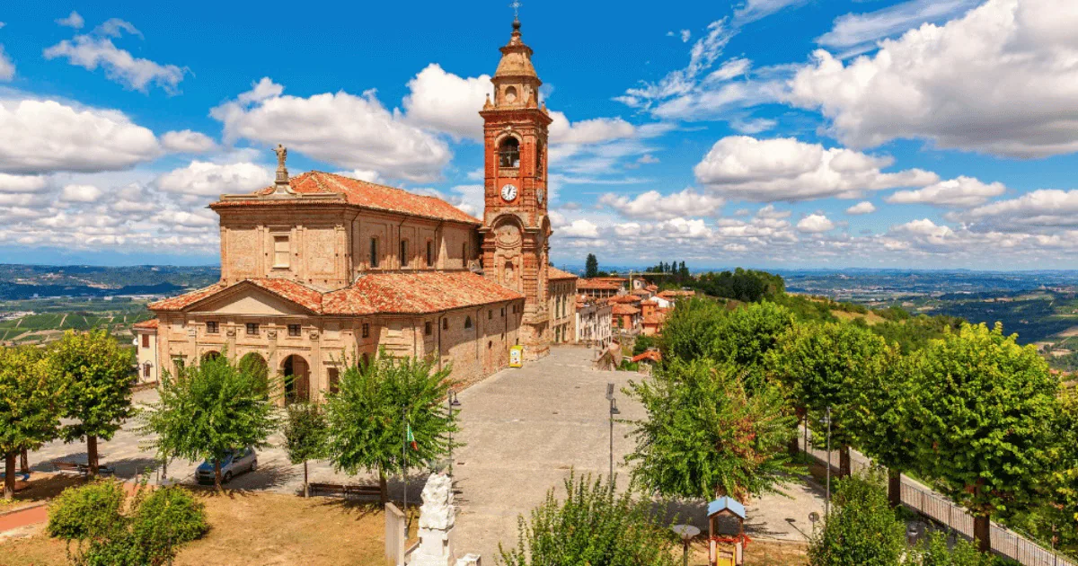 Baroque parish church with a clock bell tower overlooking the paved square of a Langhe village, vineyards and open countryside beyond.