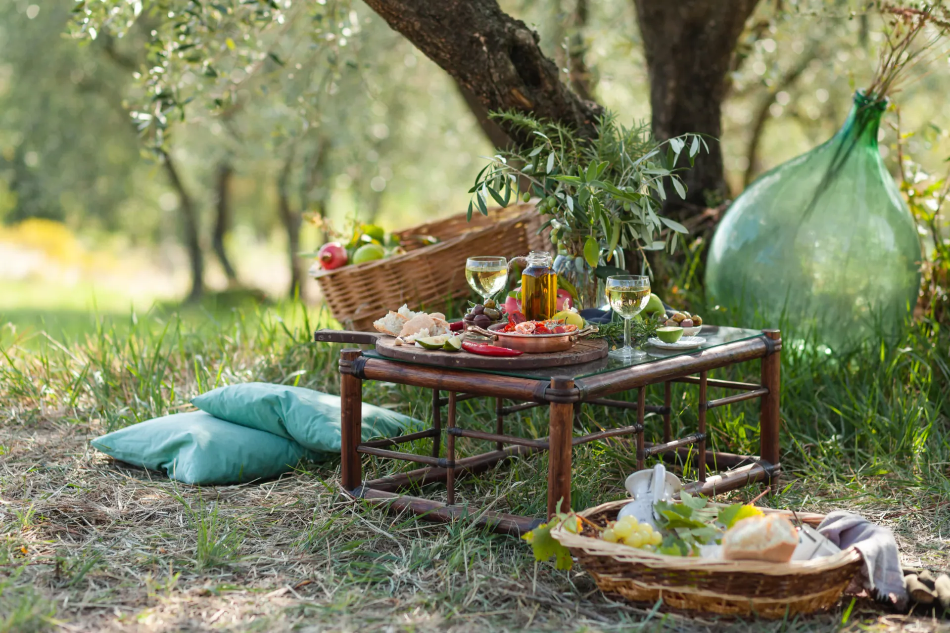 Rustic-chic picnic setting in an Italian olive grove. A low wooden table is set with white wine, fresh bread, and olive oil, surrounded by green pillows, wicker baskets, and a large vintage green glass demijohn.