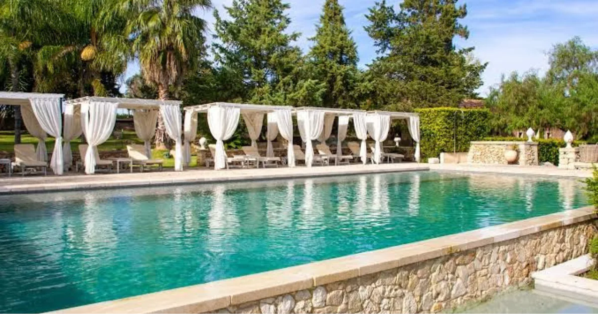 The swimming pool at Naturalis Bio Resort, lined with white curtained cabanas and sun loungers among palms and pines in the Salento countryside.