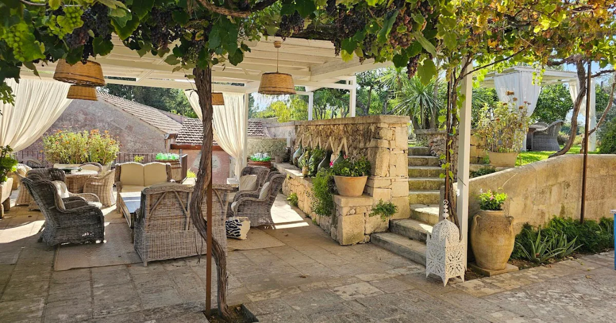 A shaded terrace at Naturalis Bio Resort under a pergola hung with ripening grapes, with wicker armchairs, stone steps and terracotta jars.