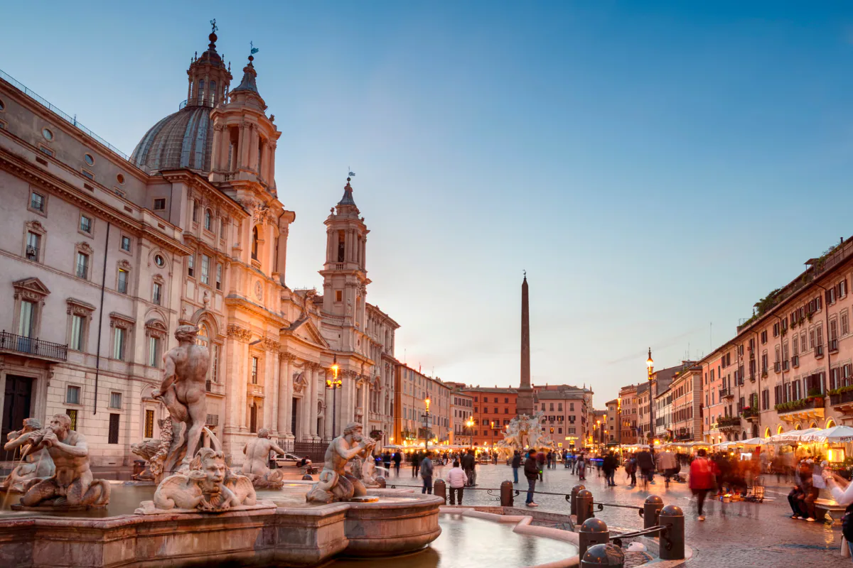 Piazza Navona at dusk, with the marble figures of its fountain in the foreground, the church of Sant'Agnese in Agone lit behind and the obelisk further down the square.