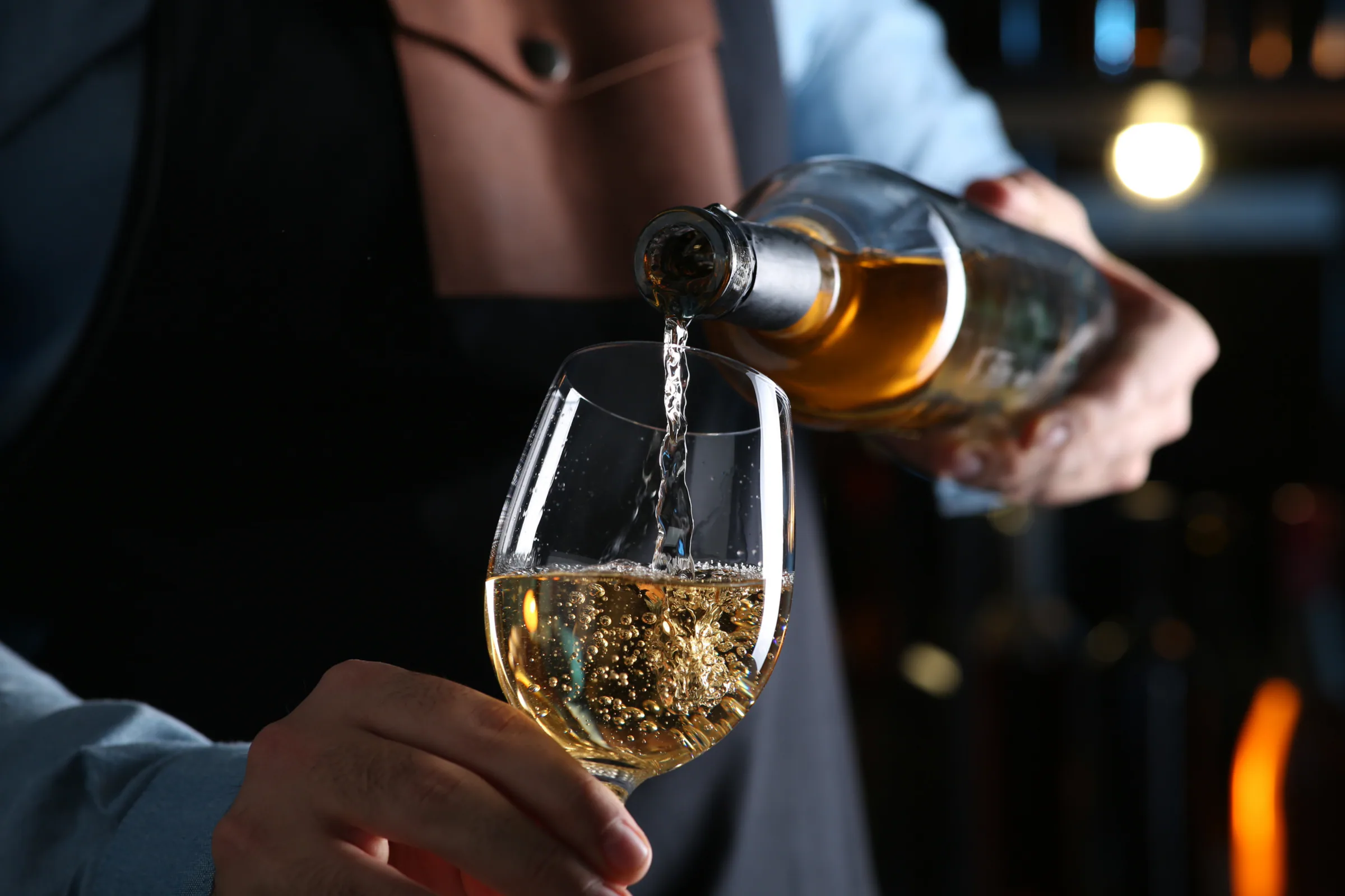 A sommelier pours white wine into a tasting glass in a dimly lit cellar.