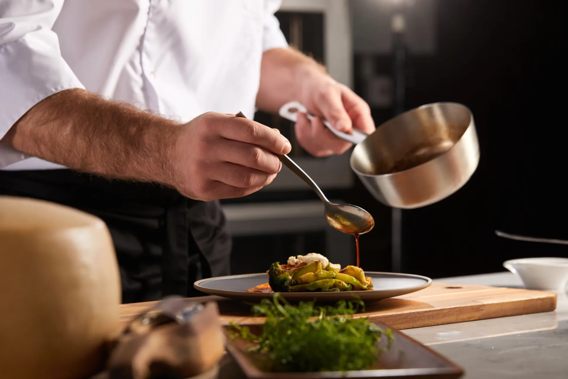 Close-up of a chef in a white uniform pouring sauce over a gourmet vegetable dish using a spoon. In the foreground, a wheel of parmesan cheese and fresh herbs sit on a wooden table, highlighting fresh Italian ingredients.