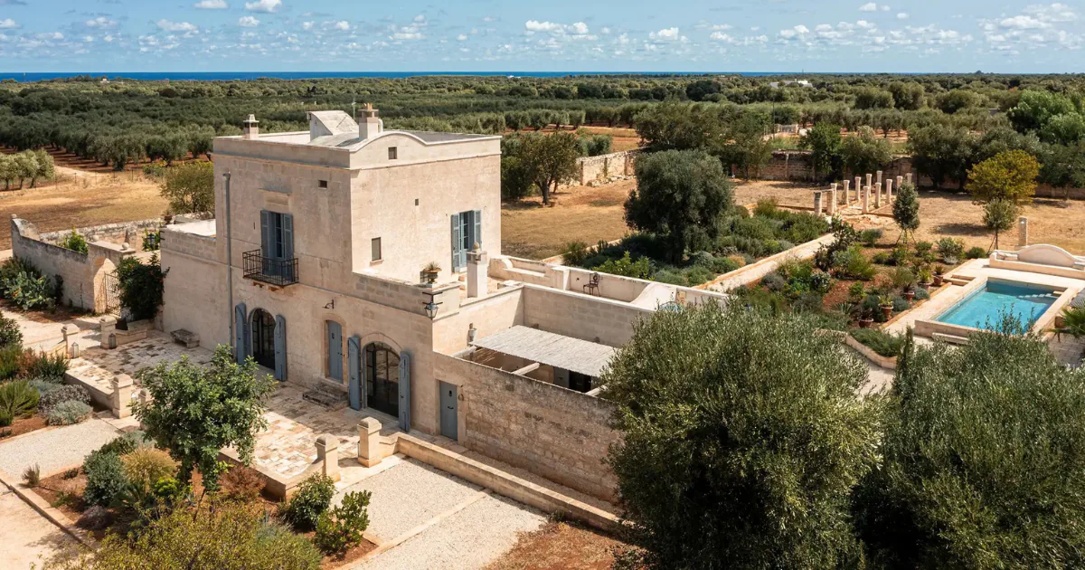 A restored stone masseria surrounded by olive groves and dry stone walls in the Salento countryside, with a swimming pool in its garden and the sea on the horizon.