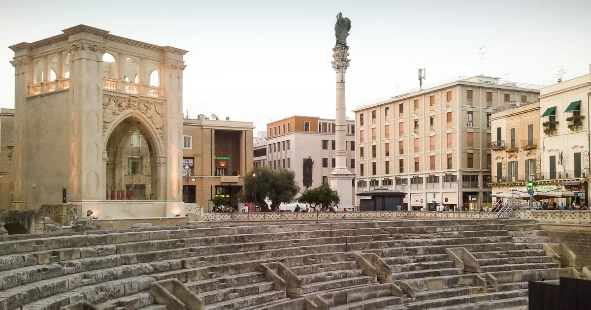 The Roman amphitheatre in Piazza Sant'Oronzo in Lecce, with the statue of the saint on its column and the city's buildings around the square.