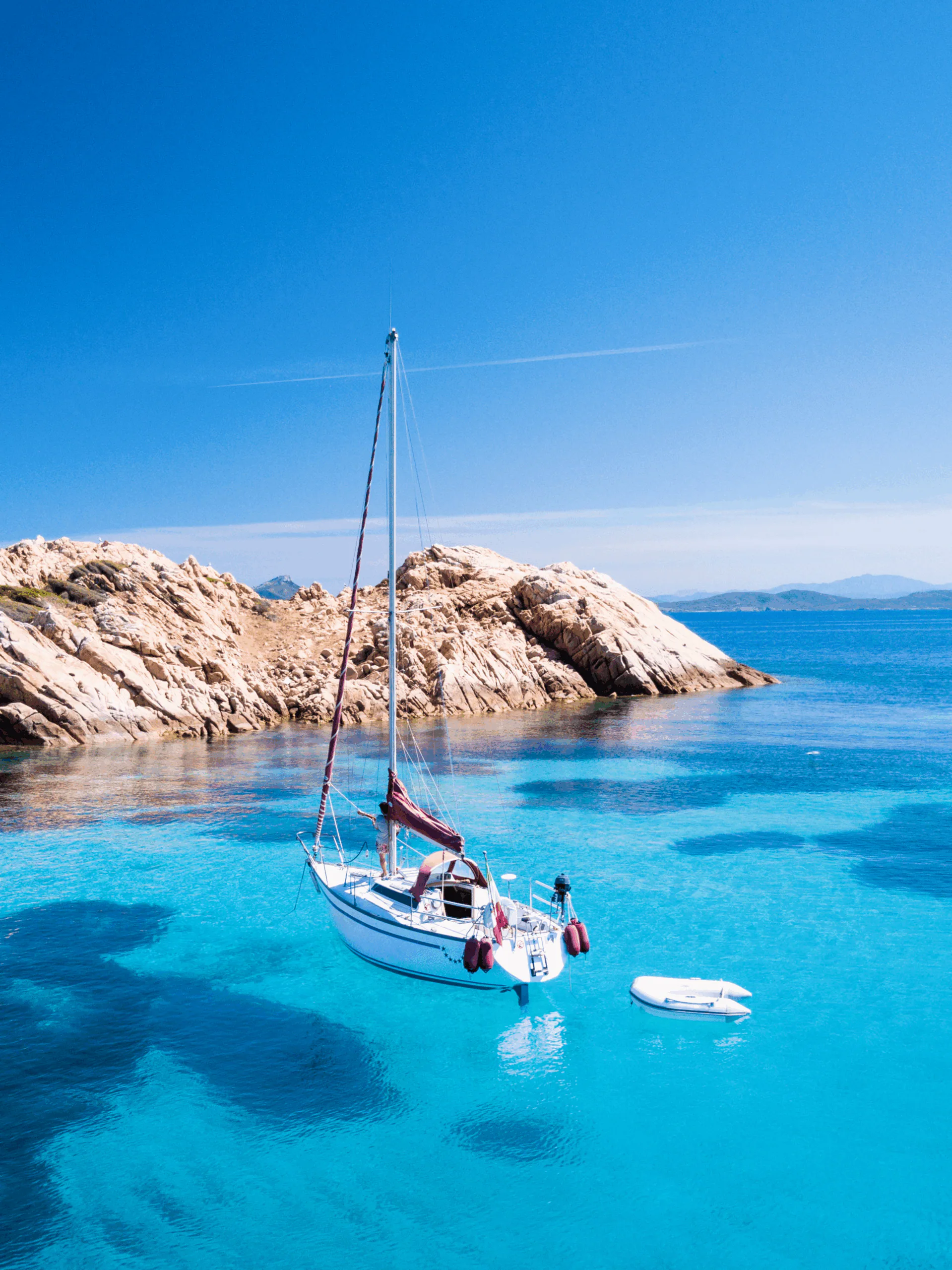 A sailing boat and its tender anchored in a turquoise cove in Sardinia, with pink granite rocks along the shore and open sea beyond.