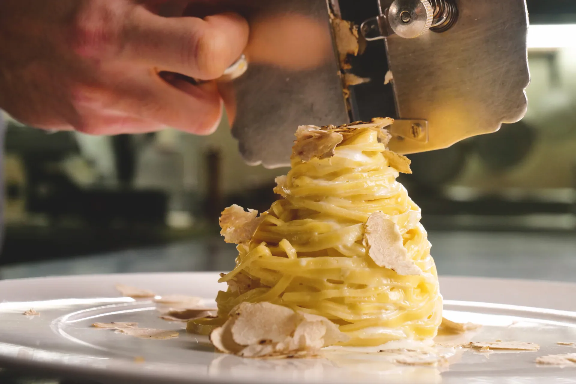 Close-up of a hand using a metal slicer to shave thin flakes of fresh white truffle over a steaming mound of buttered egg pasta (Tajarin) on a white plate.