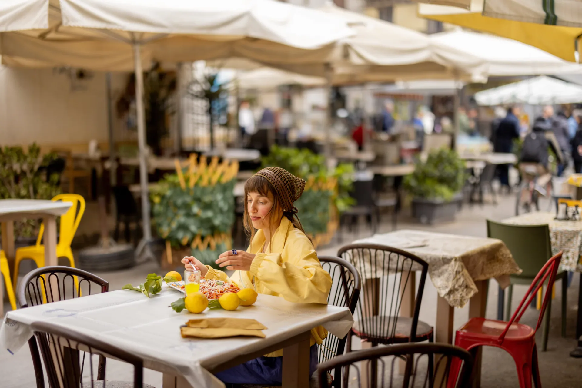 Young female tourist wearing a yellow jacket and headscarf eating pasta at an outdoor restaurant table decorated with fresh lemons. In the background, blurred white umbrellas and people in an Italian piazza, likely in Sorrento or Amalfi.