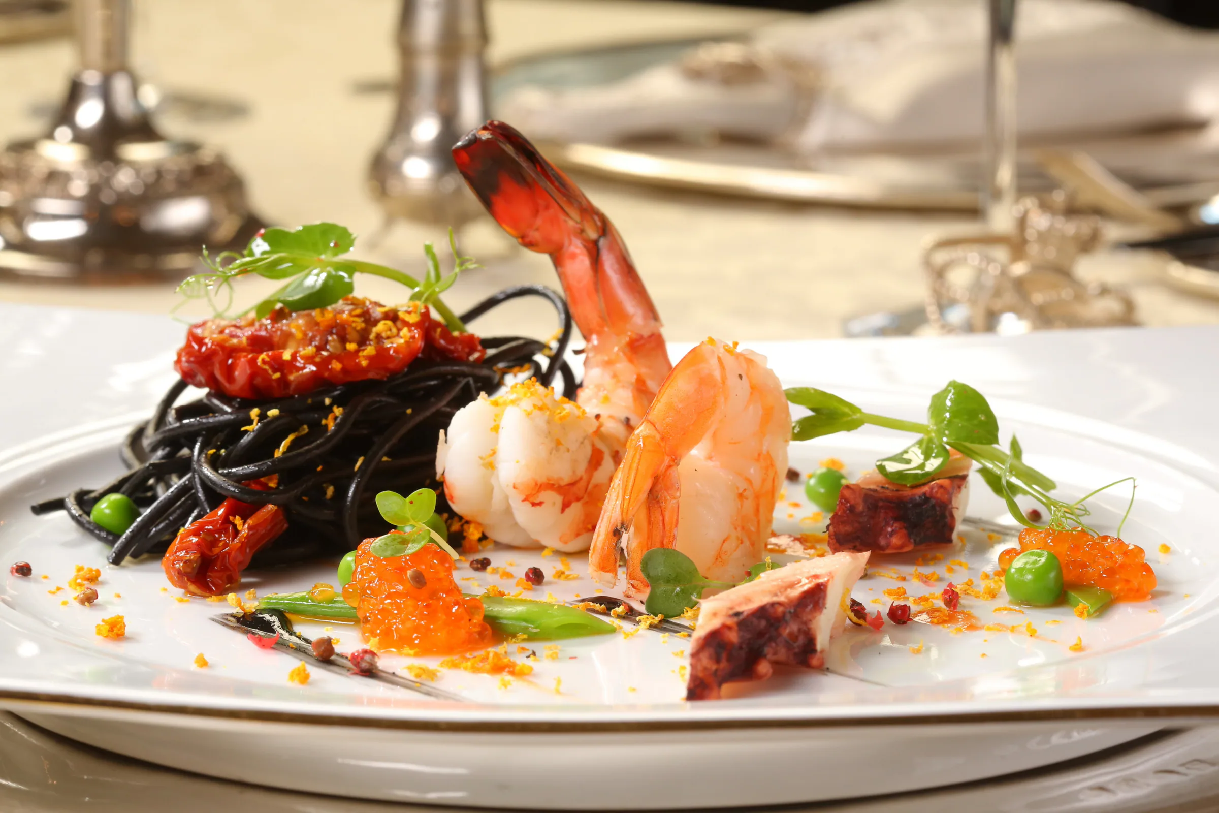 A plated dish of squid ink pasta with prawns, octopus, tomato and herbs, served on white porcelain at a formally set table.