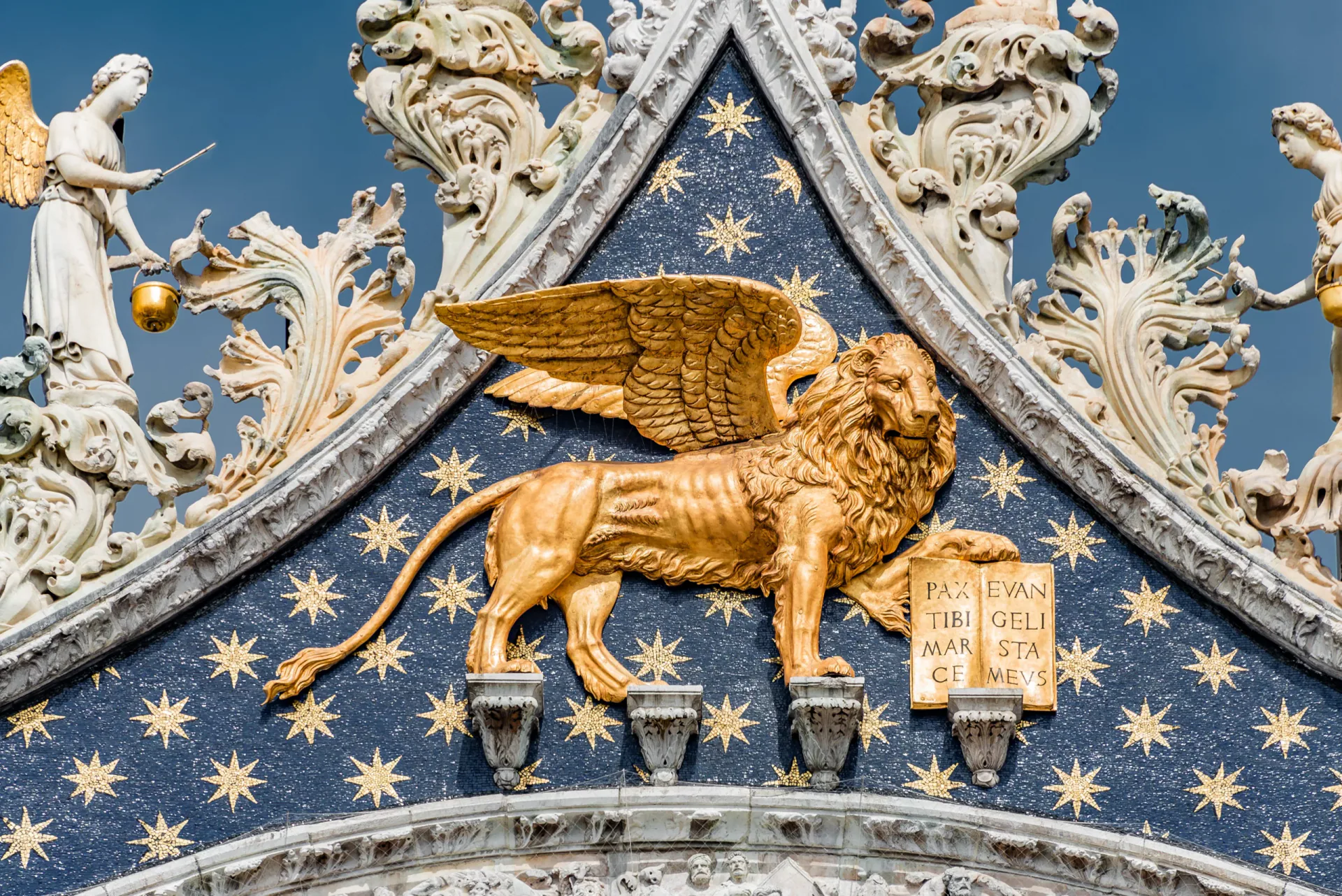 Close-up detail of the golden statue of the Winged Lion of St. Mark holding an open book against a blue starry mosaic background on the upper facade of St. Mark's Basilica in Venice.