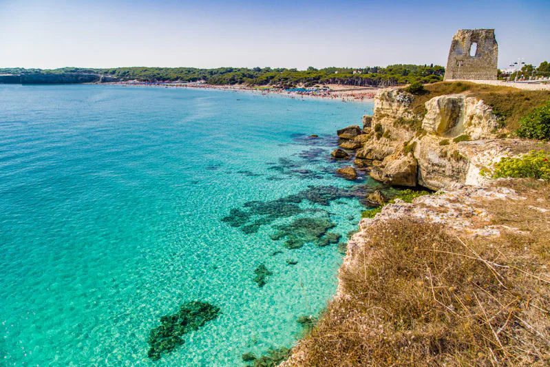 The ruined Spanish watchtower above the cliffs of Torre dell'Orso, with a turquoise bay and a sandy beach curving away below.