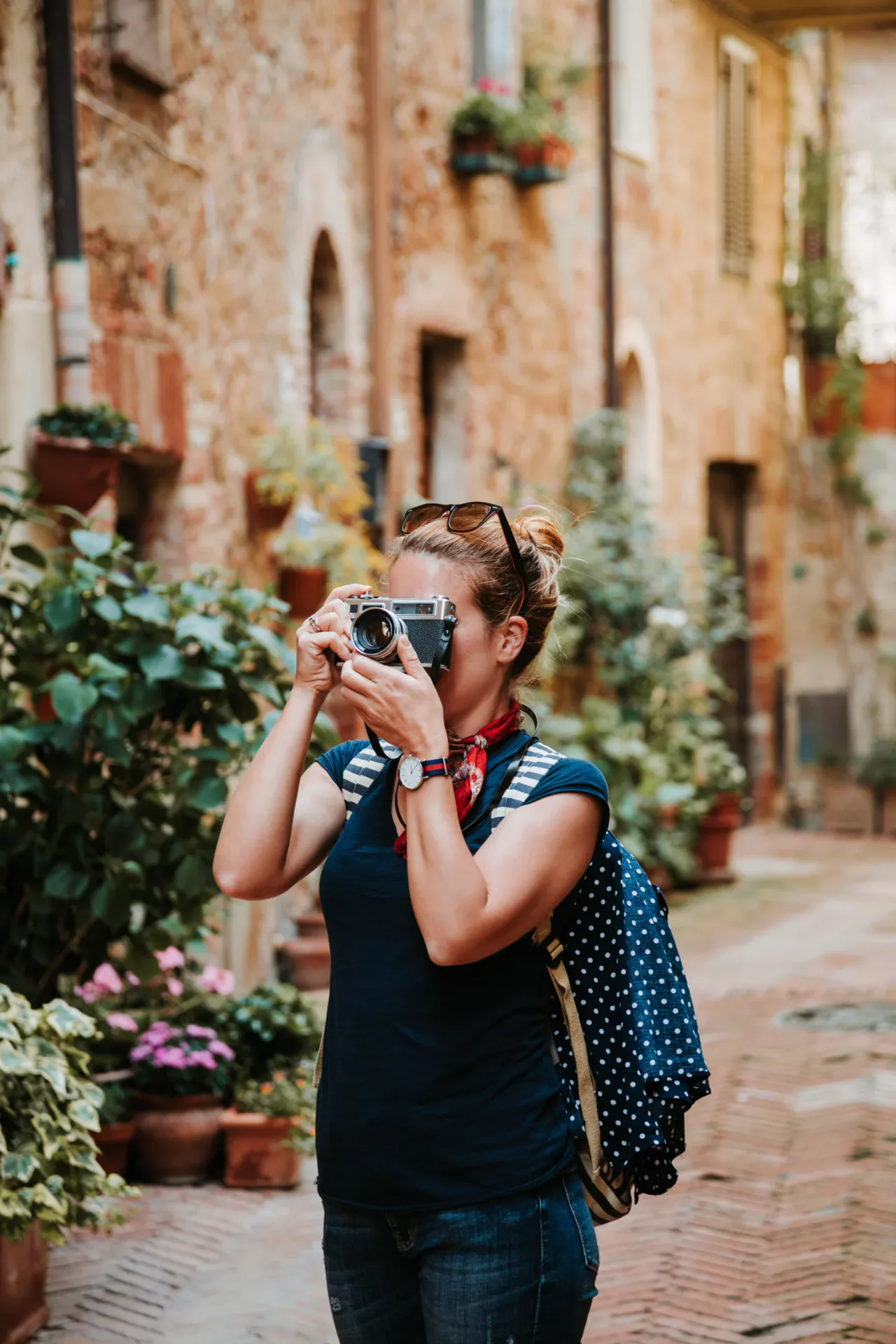 Capturing the charm: Street photography in a hidden Italian borgo alley A traveler woman with a vintage silver camera taking a photograph in a narrow, rustic cobbled alley lined with stone buildings, arches, and potted geranium flowers in a medieval Italian town.